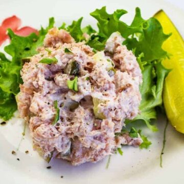 A plate with a serving of tuna salad on a bed of lettuce, garnished with a pickle spear. The salad contains chunks of tuna and appears to be mixed with mayonnaise. The plate is decorated with colorful flower designs.