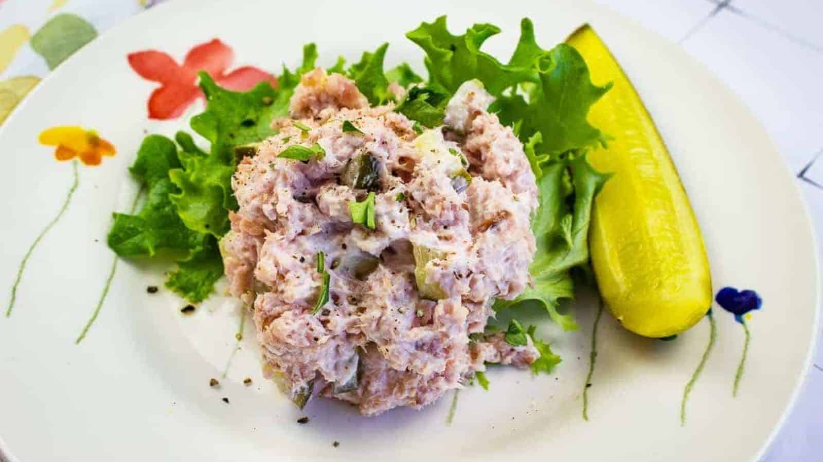 A plate with a serving of tuna salad on a bed of lettuce, garnished with a pickle spear. The salad contains chunks of tuna and appears to be mixed with mayonnaise. The plate is decorated with colorful flower designs.