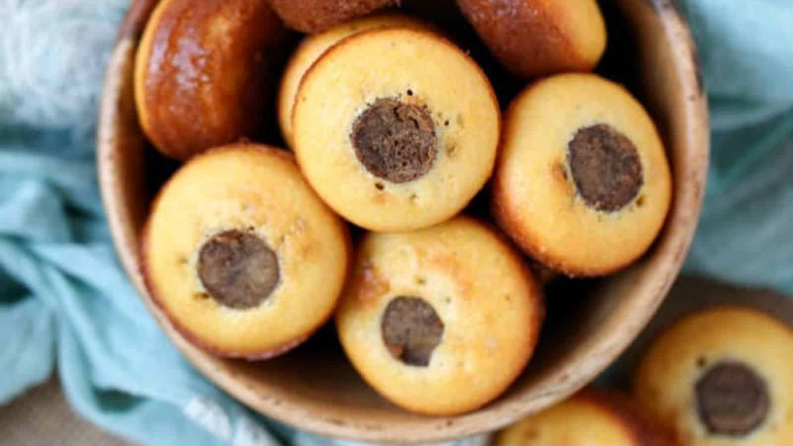 A bowl filled with cornbread muffins, each with a round sausage in the center, perfect for Kid-Friendly Recipes. The bowl sits on a surface with light blue fabric peeking around it. One muffin rests outside the bowl, sure to satisfy even the Pickiest Eaters.