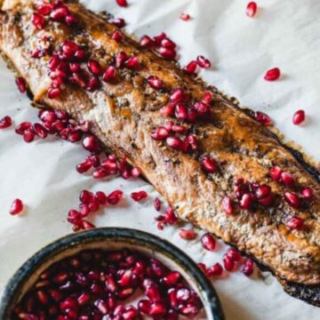 Grilled fish fillet garnished with pomegranate seeds rests elegantly on parchment paper for easy cleanup. A small bowl of extra seeds is placed beside the fillet on a rustic tray, making it an ideal dish for fans of genius recipes.
