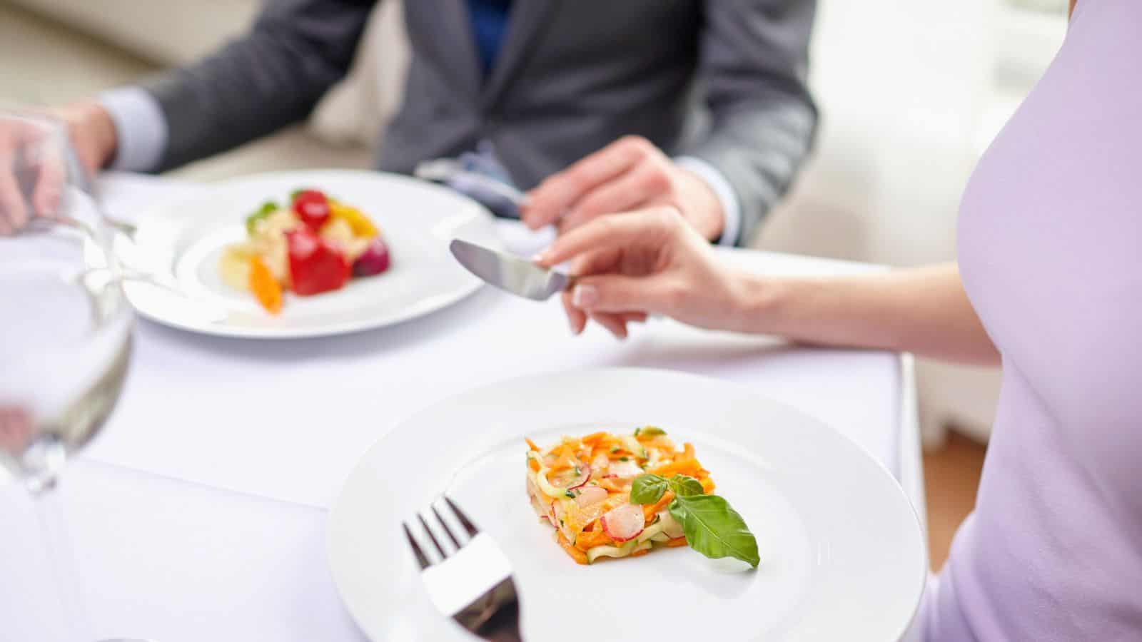 Two people are seated at a table with white linens, each with a plate of food. One plate has a vegetable dish, while the other has fruit. One person holds a knife and fork. Both are dressed in formal attire.