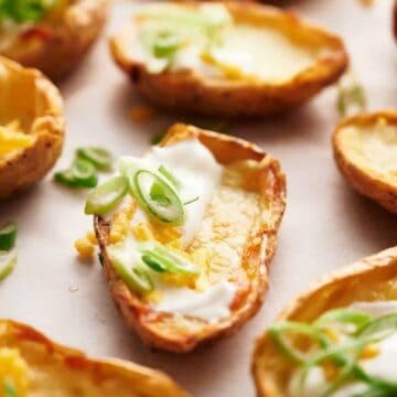 Close-up of baked potato skins topped with melted cheese, sour cream, and sliced green onions on a parchment-lined surface. The golden-brown skins are arranged closely, highlighting the toppings.