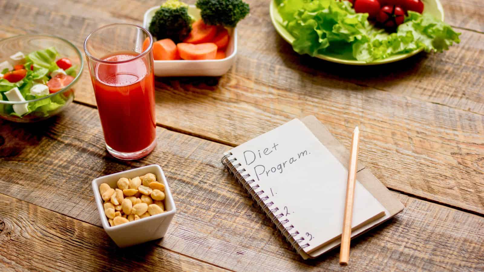 A notebook with "Diet Program" written on it lies on a wooden table with a pencil. Nearby are a glass of red juice, a bowl of peanuts, a salad, and a tray with broccoli, carrots, and cherry tomatoes.