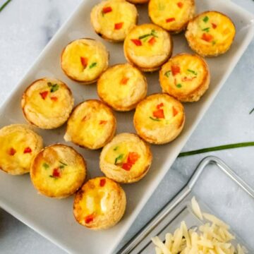 A white rectangular plate holds several mini quiches with visible bits of red and green peppers. Next to the plate is a small pile of grated cheese on a glass dish. Long green chives are scattered on a marble surface.