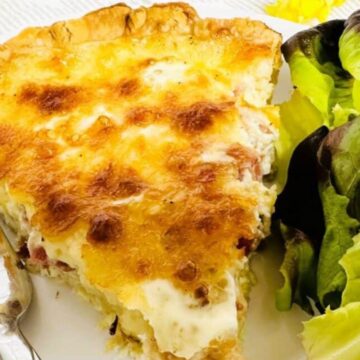 Slice of Quiche Lorraine on white plate next to fork and green salad.