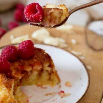 A piece of cake topped with fresh raspberries is on a white plate. A fork with a bite of cake is held above. The plate is on a wooden surface, with raspberries scattered around and a beige cloth in the background.