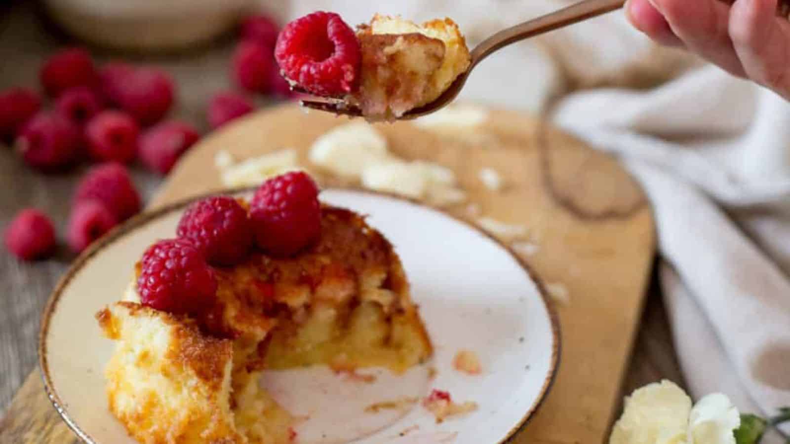 A fork holds a piece of cake above a white plate with a partially eaten slice of cake topped with raspberries. More raspberries and crumbs are scattered on a wooden board in the background.