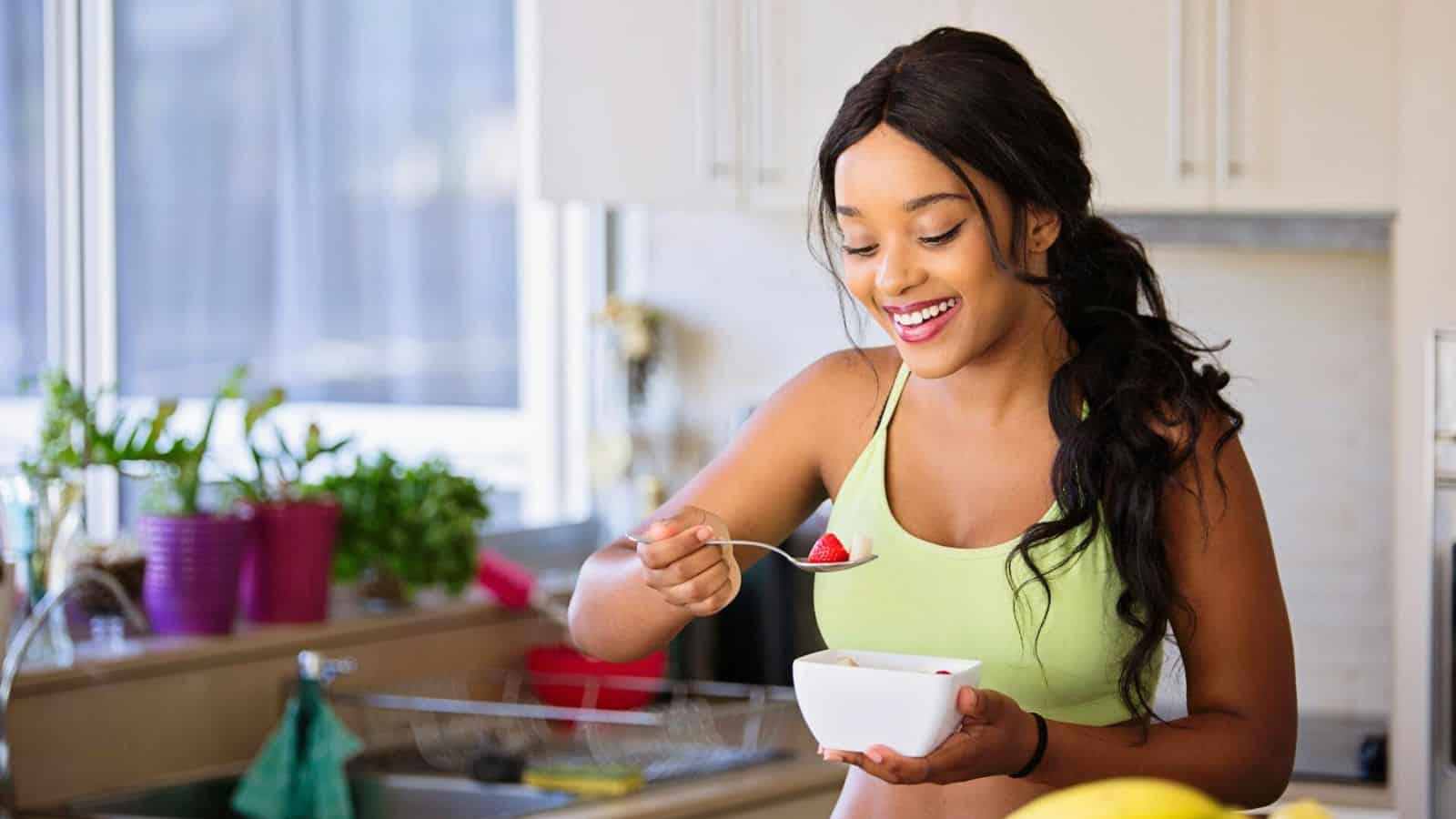 A woman in a green tank top stands in a kitchen near plants, smiling as she holds a spoon and a bowl of fruit.