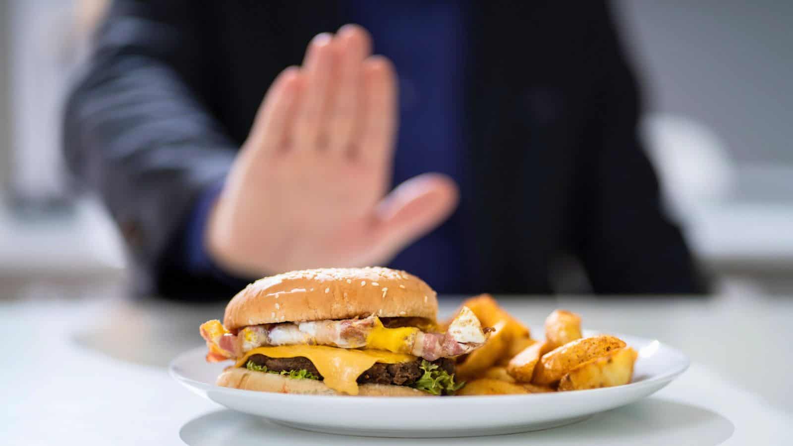 A person is gesturing to stop a hamburger on a plate in front of them. The burger is topped with cheese and bacon, and is accompanied by potato wedges. The focus is on the hand and the food.