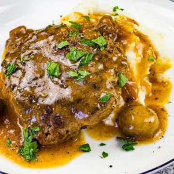 A plate of Salisbury steak with rich gravy rests on a bed of mashed potatoes, reminiscent of Grandma's comforting meals. The dish is garnished with chopped parsley and accompanied by small whole mushrooms, while a patterned napkin peeks from the background.