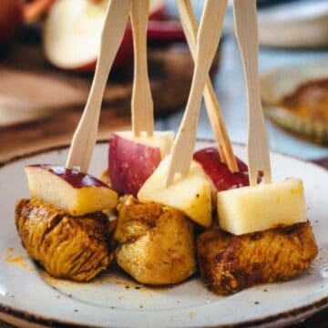 A plate with skewered pieces of cooked meat and apple cubes on wooden sticks. The dish is presented on a ceramic plate, and there is an assortment of blurred items in the background.