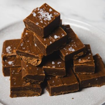 A stack of fudge squares is placed on a round, light-colored plate. The fudge is topped with coarse sea salt and situated on a marble surface.