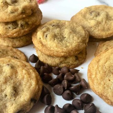 A stack of chocolate chip cookies sits on a white surface, surrounded by scattered chocolate chips. These golden brown delights with visible chocolate chunks are a true testament to why we love desserts today.