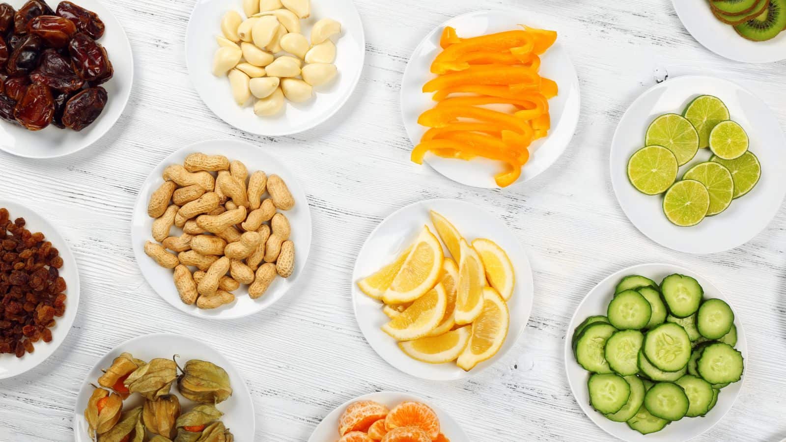 Plates with various foods including dates, garlic cloves, orange bell pepper slices, lime slices, raisins, peanuts, lemon wedges, sliced cucumber, cape gooseberries, and peeled orange segments are arranged on a white wooden table.