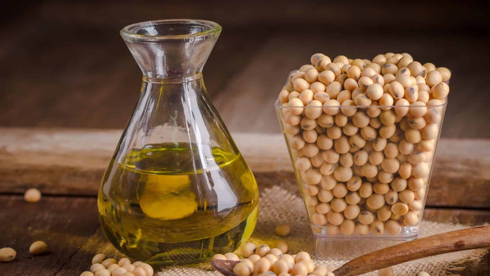 A clear glass container filled with healthy yellow oil is placed next to a square glass filled with soybeans. Both items rest on a wooden surface, with some soybeans scattered around. A small wooden spoon is partially visible in the foreground, highlighting the wholesome choice over toxic alternatives.