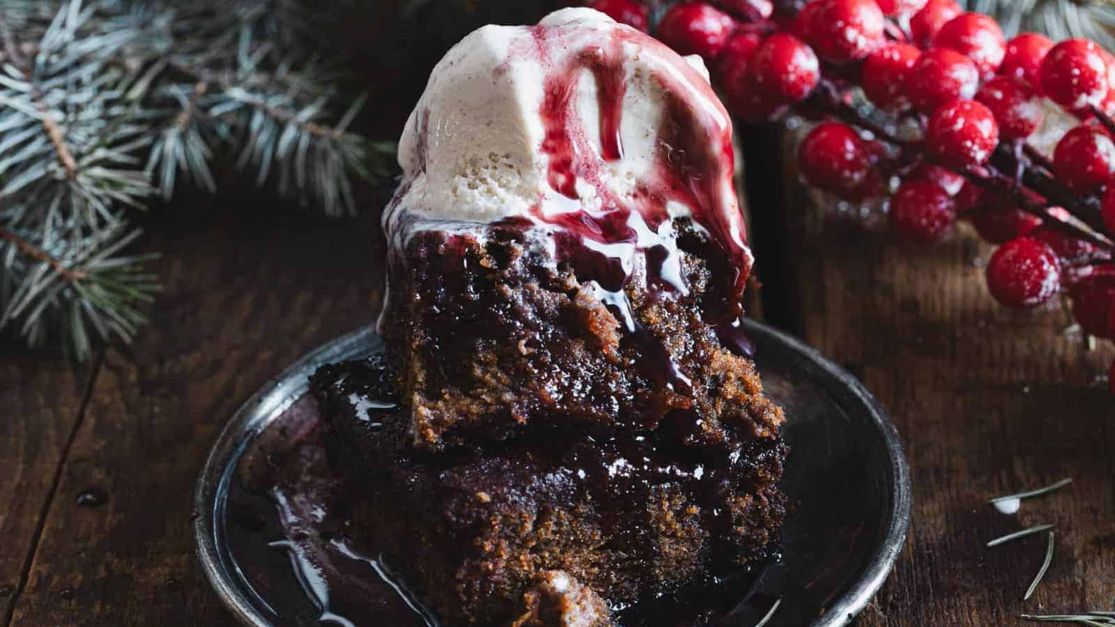 A slice of dark cake with a scoop of vanilla ice cream on top, drizzled with berry sauce, sits on a plate. Red berries and evergreen branches are in the background on a wooden surface.