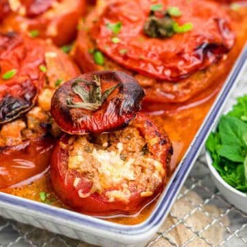 Baked stuffed tomatoes filled with minced meat and topped with their lids are arranged in a baking dish. A small white bowl with fresh parsley and oregano is placed beside the dish on a wire cooling rack.