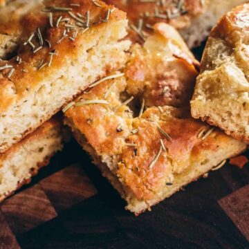 Close-up of slices of focaccia bread on a wooden surface. The bread rolls are golden brown with a fluffy texture, topped with sprigs of rosemary and coarse salt. The pieces are arranged unevenly, showcasing their airy interior and inviting you to learn the art of baking such delightful breads.