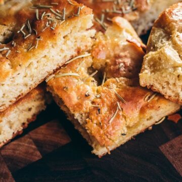 Close-up of several slices of focaccia bread on a wooden surface. The bread is golden-brown with a slightly crisp crust and is sprinkled with rosemary.
