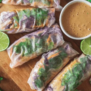 A wooden platter displays five vegetable spring rolls alongside a small bowl of brown dipping sauce. Perfect for those seeking frugal recipes, sliced limes and fresh cilantro are arranged around the platter, offering an affordable yet flavorful addition to your weekly meals.