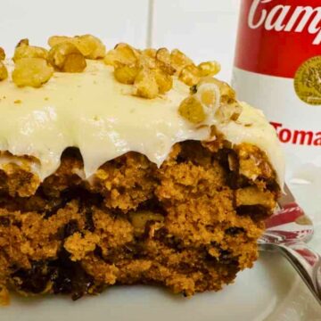 A slice of cake topped with frosting and walnuts is on a plate. In the background, there is a can of Campbell's tomato soup.