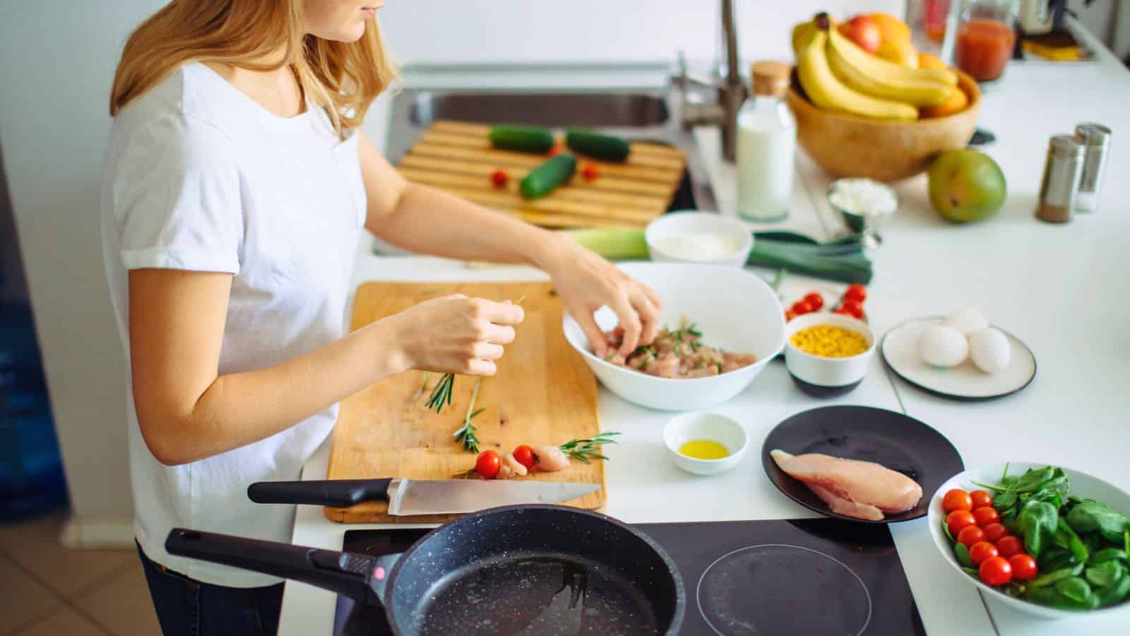 A person in a white shirt prepares food in a kitchen, arranging ingredients on a cutting board. Nearby are plates with chicken breast, cherry tomatoes, eggs, and herbs. A frying pan sits on the stovetop, and a bowl of bananas and other fruits is in the background.