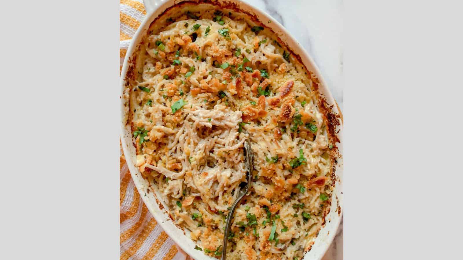 A baked casserole dish filled with a creamy pasta mixture. It is topped with golden breadcrumbs and garnished with chopped herbs. A serving spoon rests in the dish, ready for serving. A striped towel is partially visible on the side.