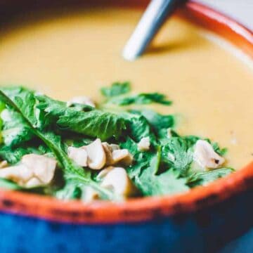 Cozy up with a close-up of a creamy yellow soup, perfect for busy days. Garnished with fresh cilantro and chopped nuts, it's served in a bowl with a blue exterior and red rim. A spoon rests invitingly in the soothing concoction.