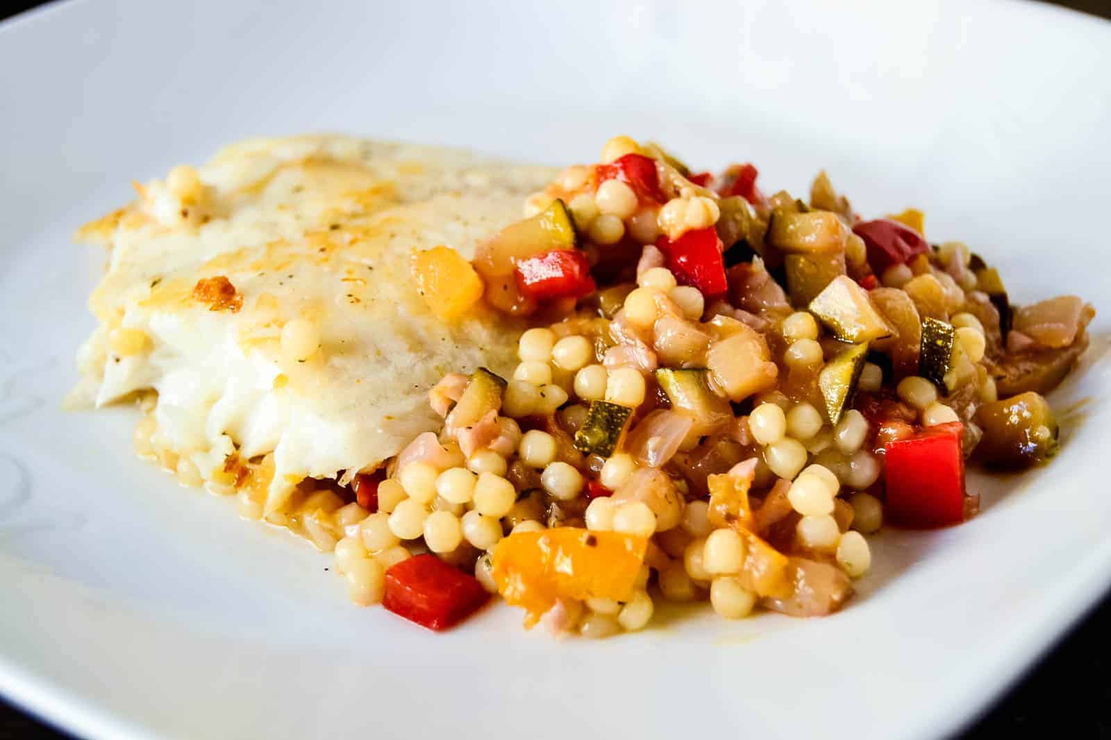 A plate with baked white fish fillet and a side of couscous mixed with diced vegetables, including red and yellow bell peppers and zucchini. This epic main course is presented on a white plate, designed to steal the show at every meal.