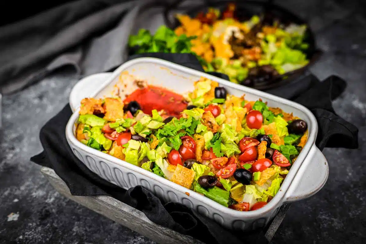 A hearty casserole dish filled with layered taco salad, featuring chopped lettuce, cherry tomatoes, black olives, and tortilla chips. The dish rests on a dark surface with a black napkin and a blurred salad in the background.