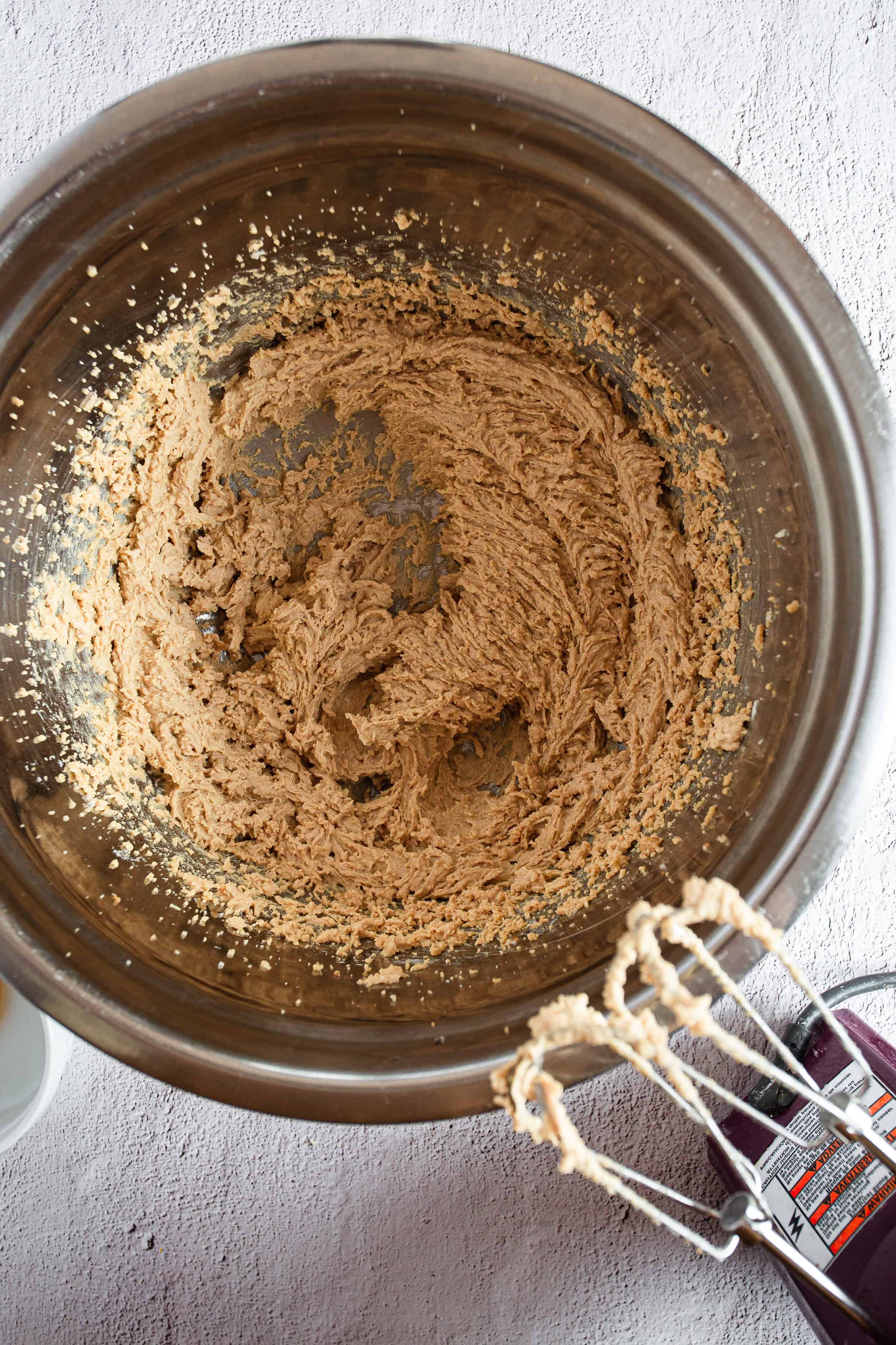 A metallic mixing bowl holds a blended mixture of gluten-free cookie dough. The dough is partially mixed, with some ingredients visibly incorporated. Electric hand mixer beaters with dough residue are visible in the foreground, ready for the addition of a Hershey Chocolate Kiss for Christmas baking fun.