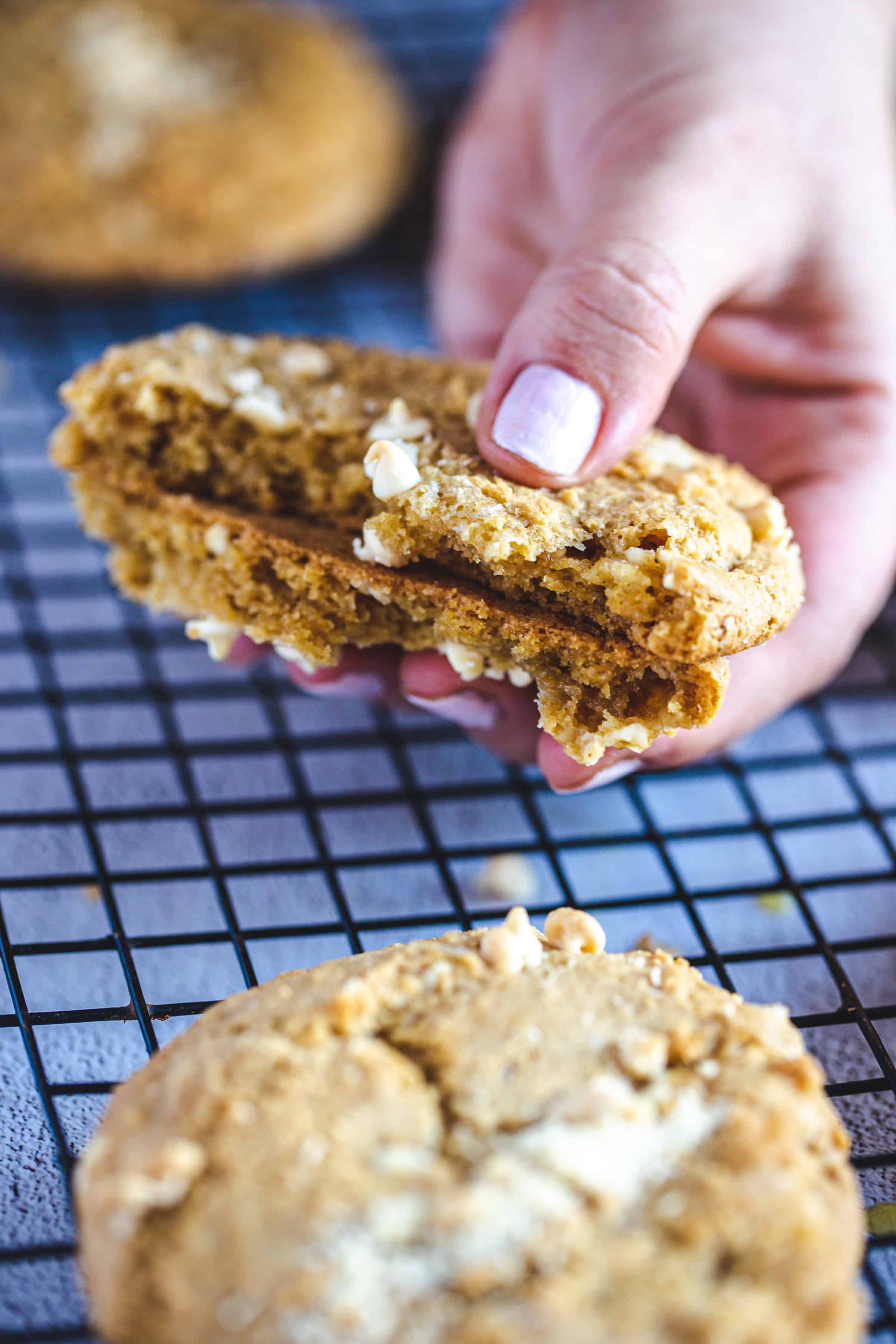 A hand is holding two halves of a gluten-free cookie with white chocolate chips, positioned over a black wire cooling rack. Another cookie rests in the background on the rack, ready to complete your Christmas cookies platter.