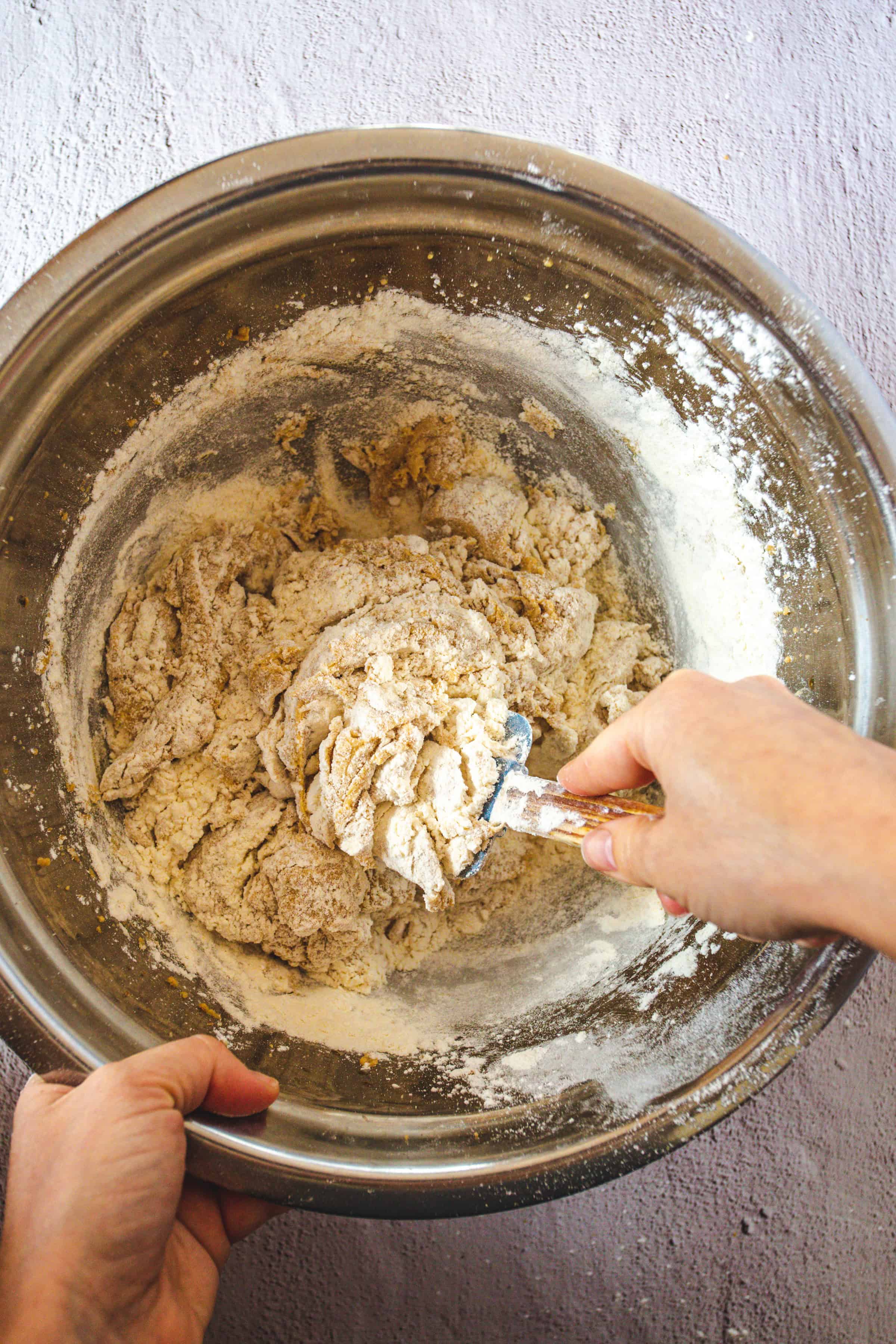 A person mixes dough for Hershey Chocolate Kiss Cookies in a large metal bowl with a spoon. The dough is partially mixed, with visible flour and clumps. The bowl rests on a textured, light-colored surface, ready to create festive Christmas treats.