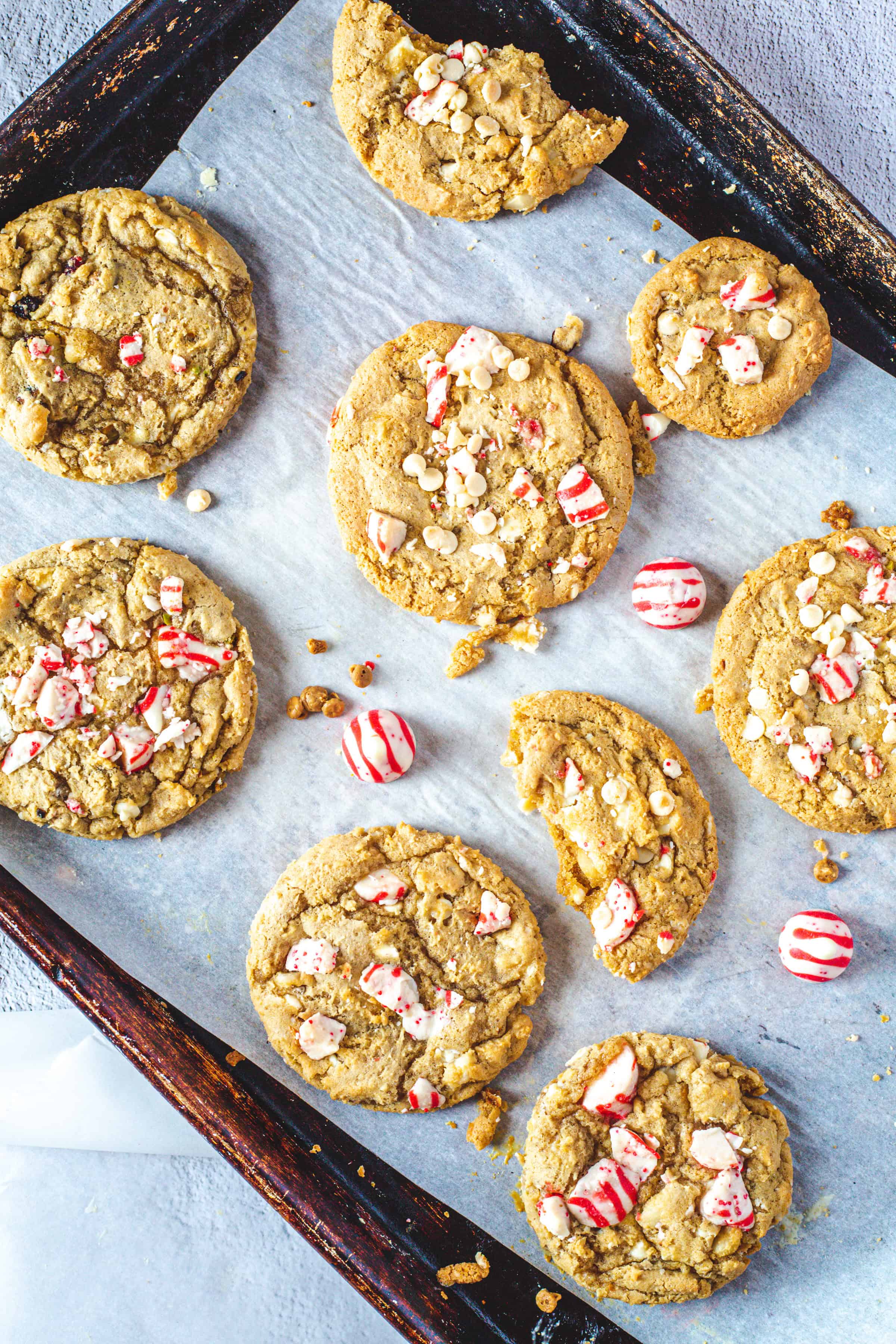 A baking tray lined with parchment paper holds several homemade Christmas cookies. Topped with crushed red and white peppermint candies, these gluten-free treats have a few scattered around. Some cookies have small bites taken out of them, capturing the sweet essence of the holiday season.