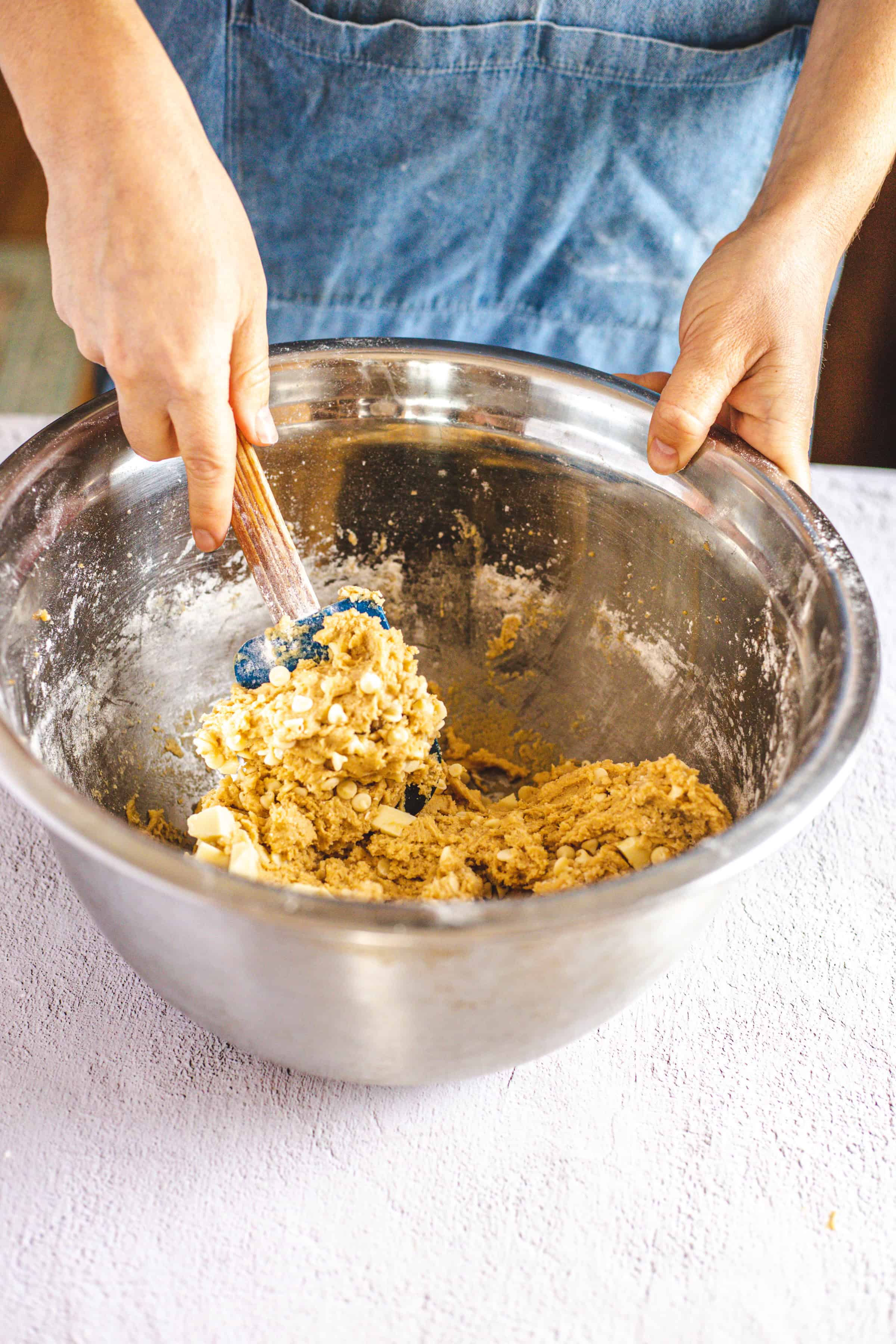 A person wearing a denim apron mixes dough for Christmas cookies in a large metal bowl with a rubber spatula on a white countertop. The dough appears to contain oats and small pieces of butter, promising festive treats.