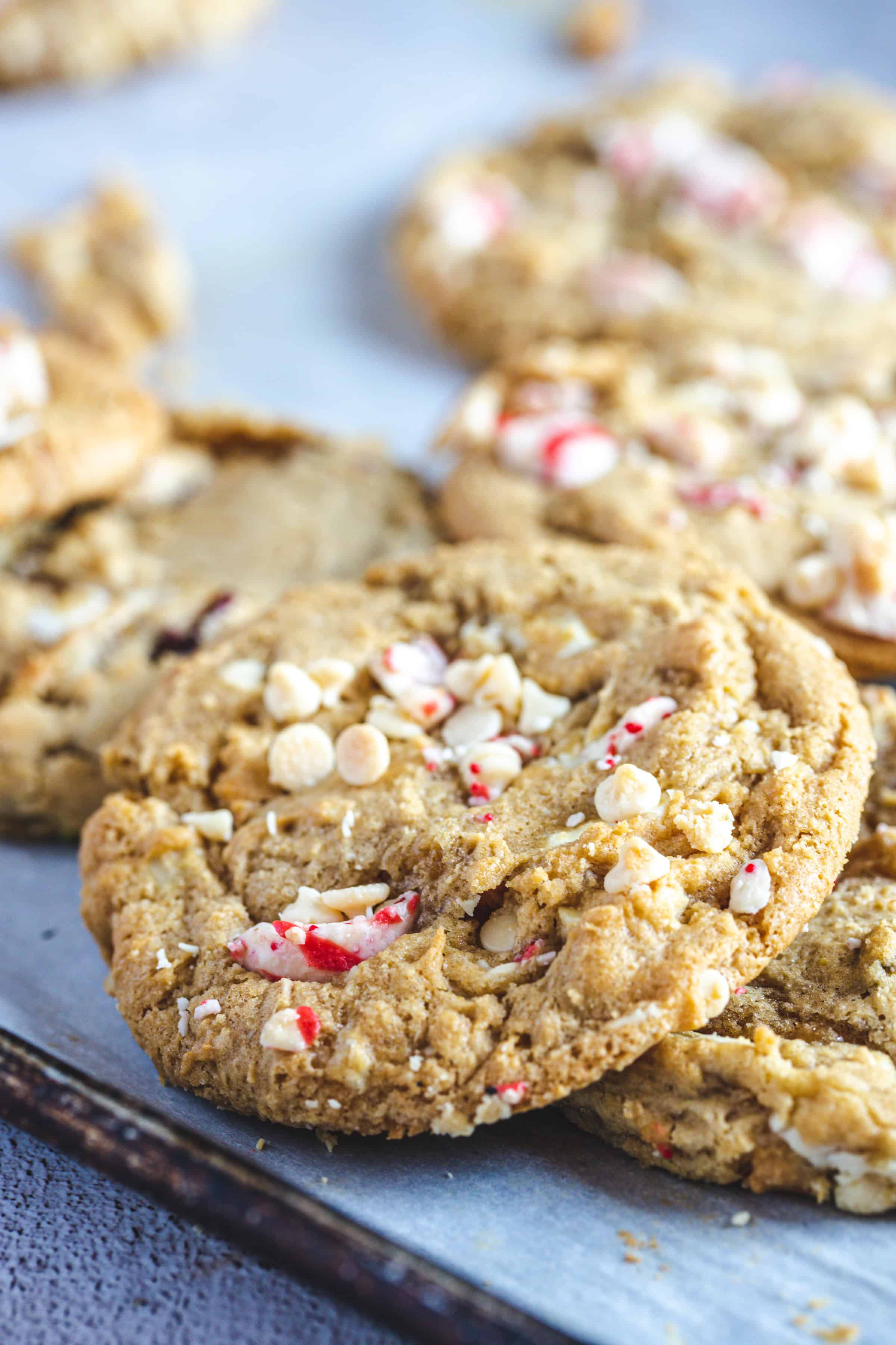 Close-up view of a batch of gluten-free cookies with visible white chocolate chips and peppermint candy pieces on a baking tray lined with parchment paper. Perfect for Christmas, they are golden brown with slightly cracked surfaces, evoking the festive spirit of Hershey Chocolate Kiss Cookies.