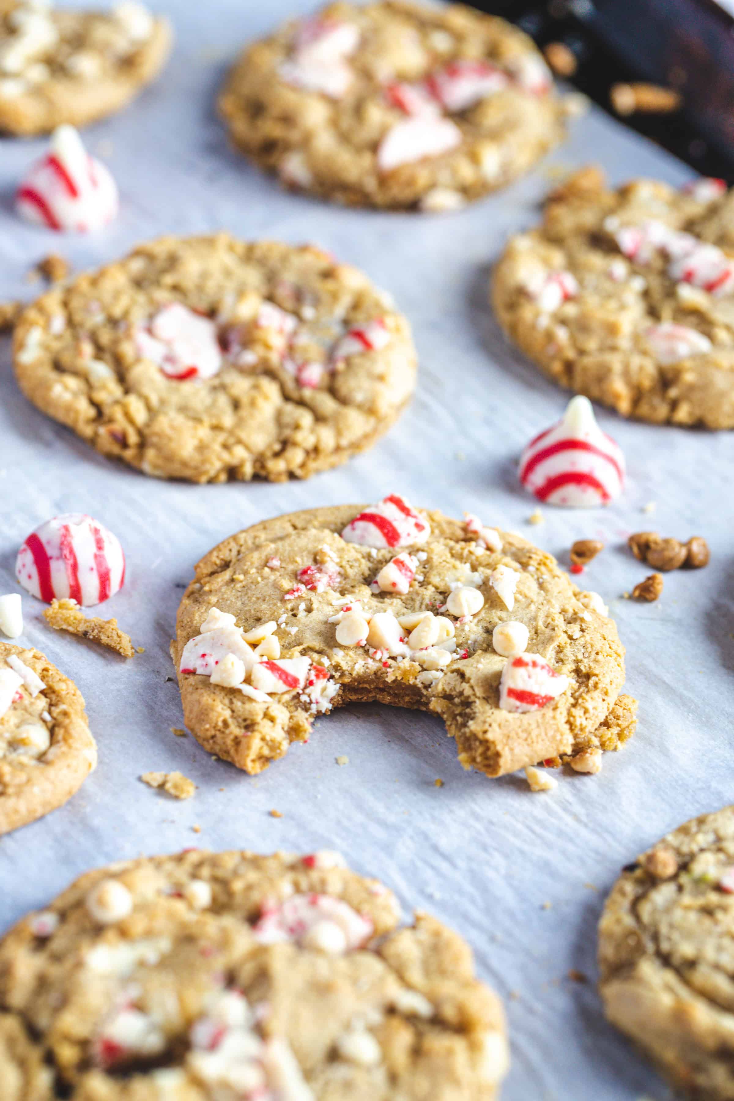 Christmas cookies topped with white chocolate chips and crushed peppermint are arranged on parchment paper. A few whole peppermint candies are also visible. One gluten-free cookie has a bite taken out of it.