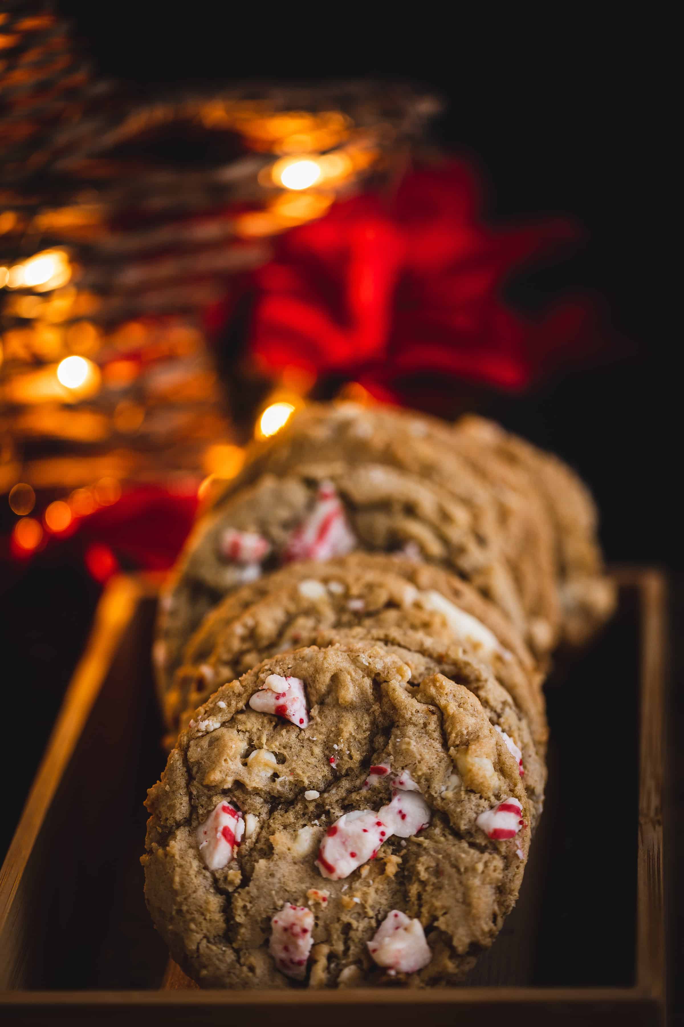 A row of delectable Christmas cookies, adorned with visible crushed peppermint pieces, sits on a wooden tray. Warm, soft lighting envelops the scene, while blurred holiday lights and red decorations add to the festive ambiance, perfect for enjoying alongside a cup of hot cocoa.