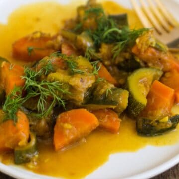 A white plate with cooked vegetables, including carrots and zucchini, garnished with fresh dill. A fork is placed on the plate's edge, and a yellow sauce covers the dish. The background is a wooden table surface.