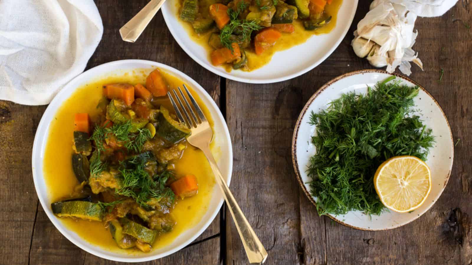 Two plates of vegetable stew with carrots and zucchini are on a wooden table. A fork rests on one plate. Fresh dill is on a small plate beside a lemon half. A bulb of garlic and a white cloth are nearby.