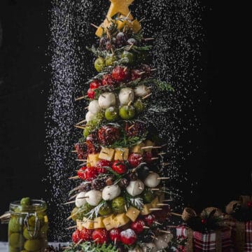 This festive Christmas tree charcuterie board doubles as a delightful antipasto display featuring skewered olives, cherry tomatoes, cheese cubes, and fresh herbs. Topped with a yellow star and sprinkled with powdered seasoning, a jar of olives sits invitingly on the side.