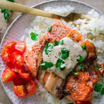 A plate of show-stopping cooked salmon topped with a creamy sauce and fresh parsley, served over white rice. Sliced cherry tomatoes are on the side. A fork rests on the plate, perfect for dinner parties. A small bowl of sauce is partially visible in the background.