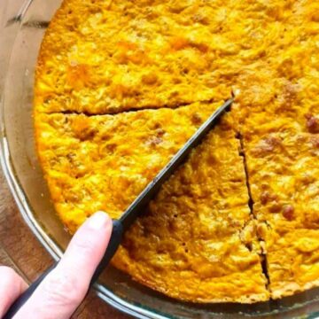 A person cuts a slice from a round, baked dish in a clear glass pie pan, reminiscent of Grandma's secret dishes. The dish boasts a golden-brown crust and rests on a wooden surface beside a few metal forks.