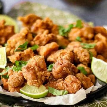 A plate of crispy fried chicken pieces garnished with lime wedges and sprinkled with fresh herbs, a true fried wonder of the world. A small bowl of sauce is on the side, placed on a patterned cloth—definitely a dish to try before you die.