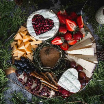 A cheese platter resembling a Christmas wreath features heart-shaped cheeses, one adorned with pomegranate seeds, surrounded by strawberries, sliced cheeses, grapes, blueberries, and crackers. The festive display rests on a wooden board with greenery entwined around it.