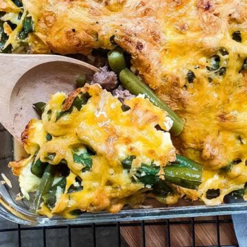 A glass baking dish filled with a classic casserole, featuring melted cheese and green beans, is perfect for any season. A wooden spoon is scooping out a portion from the corner, ready for your next potluck. The dish rests on a wire rack with "Primal Edge Health" in the corner.