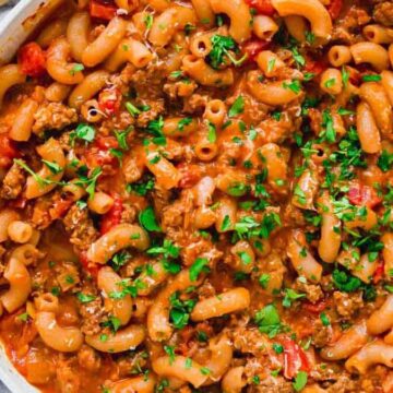 A pan filled with macaroni pasta mixed with a tomato-based sauce, ground meat, and garnished with chopped parsley. A striped napkin and wooden spoon are placed next to the pan.
