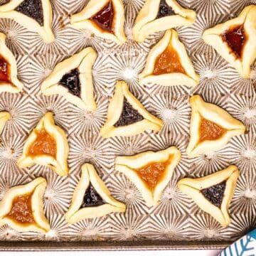 A baking tray holds several triangular pastries filled with different colored jams, including orange, red, and dark blue. The pastries are arranged in rows on the patterned tray. A blue and white patterned cloth is partially visible in the corner.