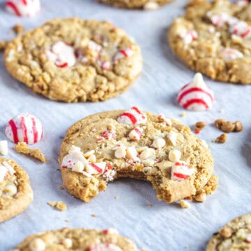 Christmas cookies with peppermint pieces and white chocolate chips sit on a parchment-lined surface. One cookie has a bite taken out of it, revealing the soft, gluten-free texture inside. Peppermint candies are scattered around, adding to the festive charm.