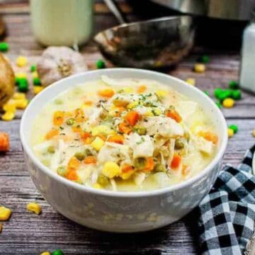 A bowl of creamy chicken soup, reminiscent of grandma's treasured recipes, sits on a wooden table. Ingredients like carrots, onions, a potato, peas, and corn surround it. There's a spoon and black-and-white checkered cloth beside the bowl, with salt and pepper shakers in the background.